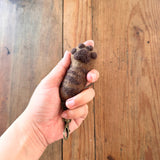 A handcrafted felt cat paw keychain is held by a female hand against a wooden background. The light brown wool felt cat paw keychain features dark brown paw pads and markings, with a textured, fur-like appearance and a copper keychain and clasp.