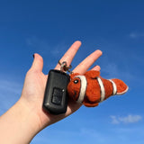 A handmade clownfish felt keychain is held by a female hand alongside a car key, set against a bright blue sky. The vibrant, high-quality plush felt clownfish features an orange body with distinctive white stripes and a small dorsal fin, attached with a metal keychain.
