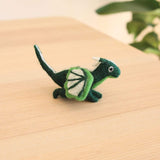 A handcrafted wool felt flying dragon keychain rests on a light brown wooden surface, complemented by a subtle backdrop of greenery. The dark green dragon features a white horn and tail, with intricately designed white and green wings. Its unique details and textures highlight its artisanal charm.