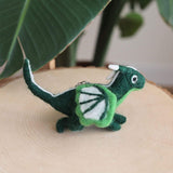 Needle felted flying dragon keychains are displayed lying on a light-colored wooden surface, set against lush green foliage. The handmade felt dragon keychains are predominantly dark green with white accents on their segmented wings, tail, belly, and horns, and feature small black eyes. A metal clasp is attached to the dragon's back.