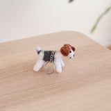 A handcrafted Jack Russell wool felt keychain rests on a light wooden surface, bathed in soft, natural light. This charming felt dog features a predominantly white body with a black back patch and a perky white tail. It has brown felt ears and black eyes, with a metal keychain and clasp attached to its back.