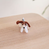 A charming handcrafted Jack Russell wool felt keychain rests on a light wooden surface. This felted keychain features a white body, brown ears, a gray and white tail, and small black eyes, with a metal clasp attached.