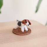 A charming Jack Russell felt keychain rests on a small, round woven coaster, which is placed on a light wooden surface with subtle greenery in the background. The handcrafted wool felt keychain features white, brown, and black felt, depicting the dog's distinctive markings, perky brown ears, and a small black nose, highlighting its artisanal appeal.