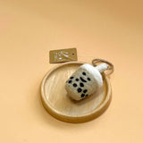 A felt Taiwanese bubble tea keychain rests on a small wooden plate against a peach-colored backdrop. The handcrafted keychain is shaped like a creamy white bubble tea cup with distinct black tapioca pearls and a white cylindrical cap. A metal keychain with a key ring is attached to the top.