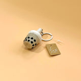 A Taiwanese bubble tea felt keychain is displayed against a peach-colored background. This handmade felt keychain features a light beige cup with visible black tapioca pearls, topped with white fluffy foam, and a metal keychain ring attached.