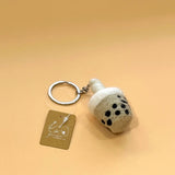 A plush felt Taiwanese bubble tea keychain rests on a peach background. This unique handcrafted felt keychain is shaped like a light grayish-beige cup, featuring black dots to represent tapioca pearls. Its rim has white plush felt, suggesting foam, and a metal keychain with a key ring is attached, from which a tag hangs.