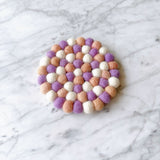 A light pink and purple round felt coaster set is displayed on a pristine marble backdrop. This handmade felt coaster set, crafted from 100% organic lamb wool, features half-inch felt balls hand-stitched in alternating light pink and purple hues, creating a soft, textured surface. The ethically handmade design, supporting female artisans, evokes a gentle warmth for home decor.