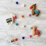 A playful wool felt llama garland is arranged on a marbled white surface. It features three 3D needle-felted llamas in shades of brown and white, each with a vibrant blue, orange, and pink striped saddle blanket and a small pink felt ball in its mouth. Colorful blue, orange, purple, and white felt balls are interspersed along the thin white string.
