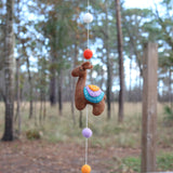 A close-up of a wool felt llama from a charming felt llama garland hangs by a white string in an outdoor forest. This skillfully needle-felted brown llama features a multi-colored saddle with vibrant orange, purple, and teal layers, enhanced by white stitch details. Colorful felt balls are interspersed along the string, adding to the garland's festive charm.