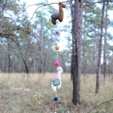 A playful wool felt llama garland is suspended outdoors from bare tree branches, with a blurred field and trees in the background. This approximately 4-foot-long garland features vibrant 3D needle-felted llamas, including one brown and one white llama, both with colorful saddles. They alternate with round felt balls in lavender, orange, pink, and navy, adding a charming, handcrafted touch.