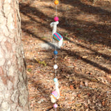 An approximately 4-foot-long wool felt Llama Garland is suspended outdoors against a tree trunk and forest floor. This vibrant garland features a string with alternating felt balls and three needle-felted, 3D llamas. The llamas are in shades of white, tan, pink, teal, and blue, while the felt balls are orange, pink, blue, and white, adding a playful touch.