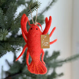 A vibrant red wool felt lobster ornament hangs from a lush green Christmas tree. The handcrafted felt lobster features detailed claws, antennae, and a jute string attached for easy hanging, adding a cheerful, nautical touch to the holiday decor.