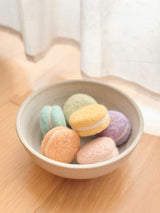 A delightful set of handcrafted wool felt macarons are artfully arranged in a light beige ceramic bowl, resting on a light brown wooden surface against a soft white curtain. These round, textured felt play food desserts come in pastel orange, pink, yellow, green, blue, and purple, each with a white filling. They are handmade with azo-free dyes, reflecting fine craftsmanship.