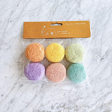 A delightful set of six handmade felt macarons, resembling classic French confections, rests on a white marble surface. These eco-friendly felt play food desserts are presented in a clear bag, showcasing their pastel colors of orange, yellow, light green, purple, pink, and aquamarine. Crafted from 100% wool and handmade in Nepal, this unique kids' toy supports fair employment for artisans.