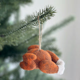 A handcrafted wool felt turkey ornament hangs against a softly blurred green and white festive background. This plump, brown turkey features textured detailing, characteristic of needle-felted craftsmanship, and distinctive white felt on its leg ends. A jute string is securely attached for hanging.