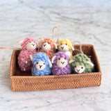 Six delightful pastel wool felt sheep ornaments are nestled in a woven rattan basket on a marbled surface. Each of these handmade felt ornaments, from a set of six, features a soft, fluffy texture and small white ears. The wool felt sheep ornaments come in charming pastel tones of pink, beige, yellow, blue, purple, and green, each with a jute string for hanging.