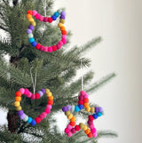 A vibrant Felt Mini Pompom Love Wreath Ornament is prominently displayed, hanging on a lush green Christmas tree. These 4-inch diameter ornaments are made from colorful 100% wool felt mini pom poms arranged in a joyful way. Suspended by a delicate jute thread, each adds a festive and handmade touch to the holiday decor.
