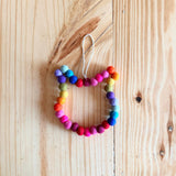 A colorful Felt Mini Pompom Cat Wreath ornament is centered on a light brown wooden surface. This circular wool felt ornament is crafted from a series of vibrant pompoms, each contributing to its textured, tactile appeal. A jute thread is attached at the top for hanging, adding to its playful, handcrafted charm.