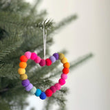 A vibrant felt Mini Pompom Love Wreath ornament hangs festively on a green Christmas tree branch. This heart-shaped wool felt ornament is crafted from small, spherical pompoms in a rainbow of red, orange, yellow, green, blue, and purple hues. A natural jute thread allows this 3D ornament to hang gracefully, standing out against a soft, blurred background.