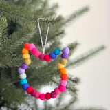 A colorful felt mini pompom cat-shaped wreath ornament hangs on a Christmas tree, creating a festive and cheerful ambiance. This cat-shaped wool felt ornament is crafted from soft, textured pompom balls in vibrant shades of red, pink, orange, yellow, green, blue, and purple. A jute thread is attached to the top for hanging.