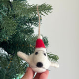 A handmade felt mini seal ornament is gently held by a female hand, positioned on a green Christmas tree, creating a cheerful holiday spirit. This charming white felt mini seal ornament features black eyes and whiskers, wearing a festive red Santa hat with a white pompom, and has a jute string for hanging.