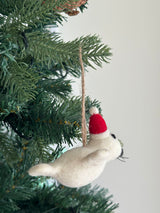 A white felt mini seal ornament, wearing a red Christmas hat with a white pompom, hangs from a lush green Christmas tree branch by a jute string. The charming handmade felt ornament features tiny black eyes and whiskers, adding a cheerful touch to holiday decor.