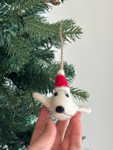 A felt mini seal Christmas ornament, wearing a red Christmas hat, is held by a female hand against a festive Christmas tree. This charming white felt seal ornament features black felt dots for eyes and nose, along with delicate whiskers. A fluffy white pompom adorns the red Christmas hat, which has a jute string attached for hanging.
