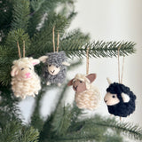 Four wool felt mini sheep ornaments are displayed on a lush green Christmas tree. These handcrafted, needle felted sheep ornaments come in white, gray, beige, and black. Each 3D sheep features small ears and a stitched mouth, suspended by a jute string, enhancing a warm holiday feel.