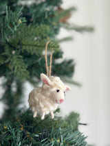 A cream-colored wool felt mini sheep ornament hangs on a green Christmas tree branch. This charming 3D felted sheep has a textured body, small pink inner ears, a pink nose, tiny black eyes, and four short legs. A jute string on its back allows for hanging, creating a festive, cozy appeal against a blurred white background.