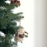A charming 3D needle-felted mini sheep ornament hangs from a Christmas tree branch, creating a cozy and festive mood. This unique Christmas ornament features a cream-colored, fluffy body crafted from organic wool, with dark brown for its face, ears, and legs. It is suspended by a jute string, adding a rustic touch perfect for holiday decoration.
