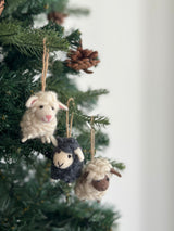 Felt mini sheep ornaments adorn a festive Christmas tree, creating a joyful atmosphere. These adorable 3D wool felt sheep ornaments are seen in various colors, including white with a pink nose, dark gray, and off-white with a brown face, each needle felted and hand-stitched from organic wool.
