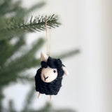 A cute wool felt mini sheep Christmas ornament hangs from an evergreen branch. This handcrafted 3D sheep ornament features a fluffy black body and head, contrasting with a smooth cream face and inner ears. Its endearing expression is formed by minimalist black stitched eyes and a small V-shaped mouth. Tiny white legs dangle below, and a jute string is attached for hanging.