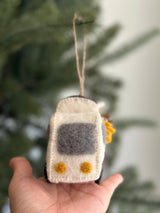 A handmade felt mobile flower shop Christmas ornament is displayed against a blurred Christmas tree background. This unique felt ornament features a white truck with a gray window, overflowing with vibrant sunflowers. A jute string is attached to the top for hanging.