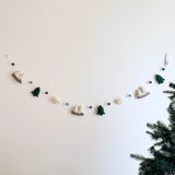 A playful wool felt mountain and cloud garland hangs against a bright white wall, featuring nature-inspired 3D designed felt figures. This unique garland showcases two-tone gray and white mountains, deep green trees, and white clouds, all connected by string and interspersed with felt balls. The handmade felt garland is secured by a transparent hook, with a green Christmas tree visible in the lower right.