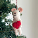 A charming wool felt mouse holding heart ornament is nestled among the green branches of a Christmas tree. This white mouse features light pink ears, tiny black eyes, and a simple black Y-shaped face. It clutches a bright red heart, with a red and white string attached for hanging.