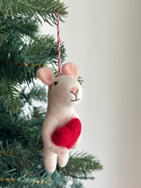 A festive handmade felt mouse Christmas ornament, holding a red heart, hangs from a green Christmas tree branch against an off-white background. This white, 3D needle-felted mouse features light pink inner ears and a stitched black nose and mouth. A red and white cotton string is attached for hanging.