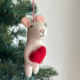 A festive handmade felt mouse Christmas ornament, holding a red heart, hangs from a green Christmas tree branch against an off-white background. This white, 3D needle-felted mouse features light pink inner ears and a stitched black nose and mouth. A red and white cotton string is attached for hanging.