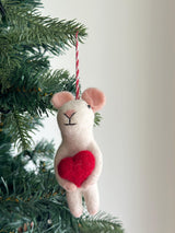 A charming Felt Mouse Holding Heart Ornament, handcrafted from wool felt, hangs on a festive Christmas tree against a soft white background. This creamy white mouse felt ornament features small, rounded pink ears, a stitched face with a 'Y'-shaped nose and black eyes, and holds a vibrant red heart. A red and white cotton string is attached to its head for hanging.