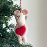 A charming Felt Mouse Holding Heart Ornament, handcrafted from wool felt, hangs on a festive Christmas tree against a soft white background. This creamy white mouse felt ornament features small, rounded pink ears, a stitched face with a 'Y'-shaped nose and black eyes, and holds a vibrant red heart. A red and white cotton string is attached to its head for hanging.