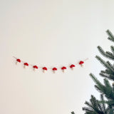 A felt mushroom garland is displayed against a plain white wall next to a lush green pine tree branch. This charming felt garland features several evenly spaced felt mushrooms, each with a vibrant red cap dotted with white spots and a soft, white stem. The mushrooms are strung on a thin, light brown string, attached to the wall with clear fasteners, evoking a whimsical natural scene.