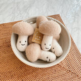 Multiple felt mushroom keychains are charmingly displayed in an off-white bowl resting on a woven mat, with a brand tag visible among them. Each handmade felt keychain features a rounded light brown cap and a white stem adorned with a sweet smiley face, topped with a metal ring. Their soft, felted texture enhances their cheerful, handcrafted appeal, creating a cozy and abundant scene.
