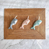 Three Felt Narwhal Ornaments are playfully displayed on a rectangular woven rattan placemat, set against a smooth marble backdrop. Each unique handcrafted felt narwhal features a distinct white horn and small white body dots, with jute strings for hanging. They come in charming light blue, pink, and green colors.