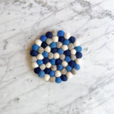 A handcrafted navy blue round felt coaster is displayed on a bright white marble surface, showcasing its calm, minimalist aesthetic. This soft, textured wool felt coaster is meticulously formed from a dense arrangement of hand-stitched navy blue felt balls made from 100% organic lamb wool, evoking a sense of warmth and handcrafted charm.