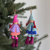 Two felt Nordic girl ornaments hang festively on a Christmas tree, each with distinct colors and textures. One ornament, dressed in a pink pointy hat, light purple coat, and pink star-embroidered dress with fuchsia boots, hangs beside another wearing a light blue hat, light blue coat, and dark blue star-embroidered dress with fuchsia boots. They are suspended by jute strings, adding a rustic, whimsical touch.