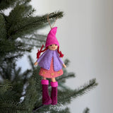 A felt Nordic girl ornament hangs on a green pine tree branch, set against a soft, blurred white background. This handcrafted wool felt ornament features a pink pointed hat, a lavender coat, and magenta boots, suspended by a brown jute string. It creates a joyful and festive holiday display.