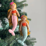 Two assorted wool felt mermaid ornaments hang from brown jute strings on a Christmas tree, set against a soft grey background. These handmade felt mermaids feature tan faces, stitched eyes, red smiles, and long golden yarn hair adorned with unique felt flowers. One has a pink beaded tail and orange seashell top, while the other has a light blue beaded tail and orange seashell top. Both have bendable arms and tails for posing.
