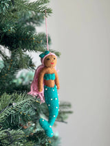 A handmade wool felt mermaid ornament hangs from a pink string on an evergreen Christmas tree, set against a soft, ambient background. The mermaid has a brown body, a turquoise seashell bra, and a matching turquoise tail with white dots. Light pink hair and a green bow on her head complete the whimsical design.