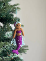A handmade wool felt mermaid ornament hangs from a Christmas tree branch by a light blue string. This unique Christmas felt mermaid ornament features a warm brown skin tone, a purple top, and a purple tail with white dots. Her light blue hair is styled with a matching cap. The felt mermaid's bendable arms and tail add a whimsical touch to holiday decor.