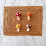 Autumn Tone Mushroom Set of 4 felt ornaments rest on a textured woven mat against a bright marble surface. Each handcrafted wool felt mushroom ornament features a charming toadstool design with a rounded, white polka-dotted cap in warm red, orange, yellow, or brown, and a cylindrical stem. A jute string tops each unique Christmas ornament, creating a natural, inviting display.