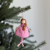 A delicate felt ballerina ornament is suspended by a natural jute string on a Christmas tree, set against a soft, light backdrop. This hand-felted wool ballerina features a soft pink wool felt dress, light peach upper body and legs. A brown felted braid adorned with small white beads completes the charming and whimsical design.